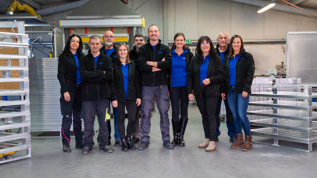 Team von Hercher Metallbau in der Produktionshalle – Gruppenfoto der Mitarbeitenden mit blauen Firmenpoloshirts und Arbeitskleidung.
